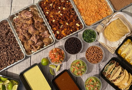 A catering spread featuring a tray of tacos, a tray of taco shells, and a chafing dish of enchiladas on an outdoor table.