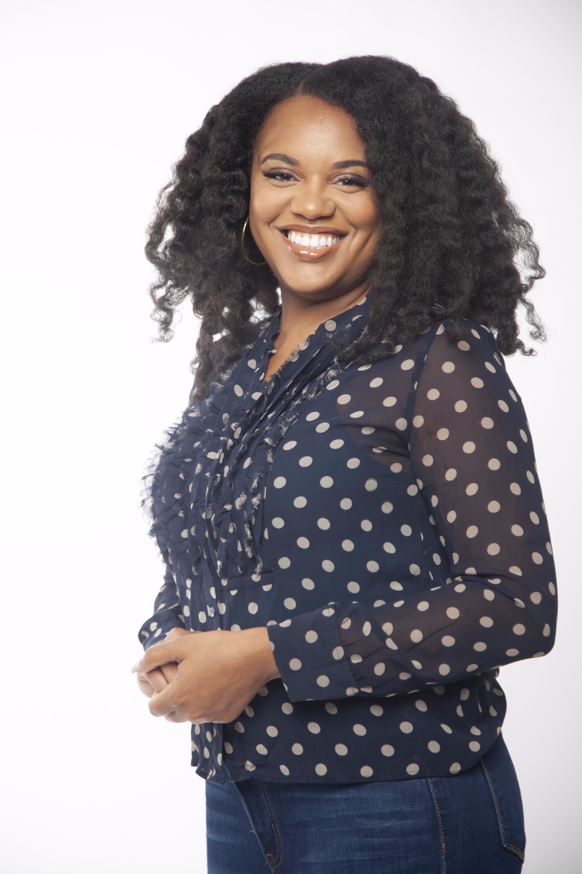 A woman with curly hair is wearing a blue polka dot shirt and jeans.