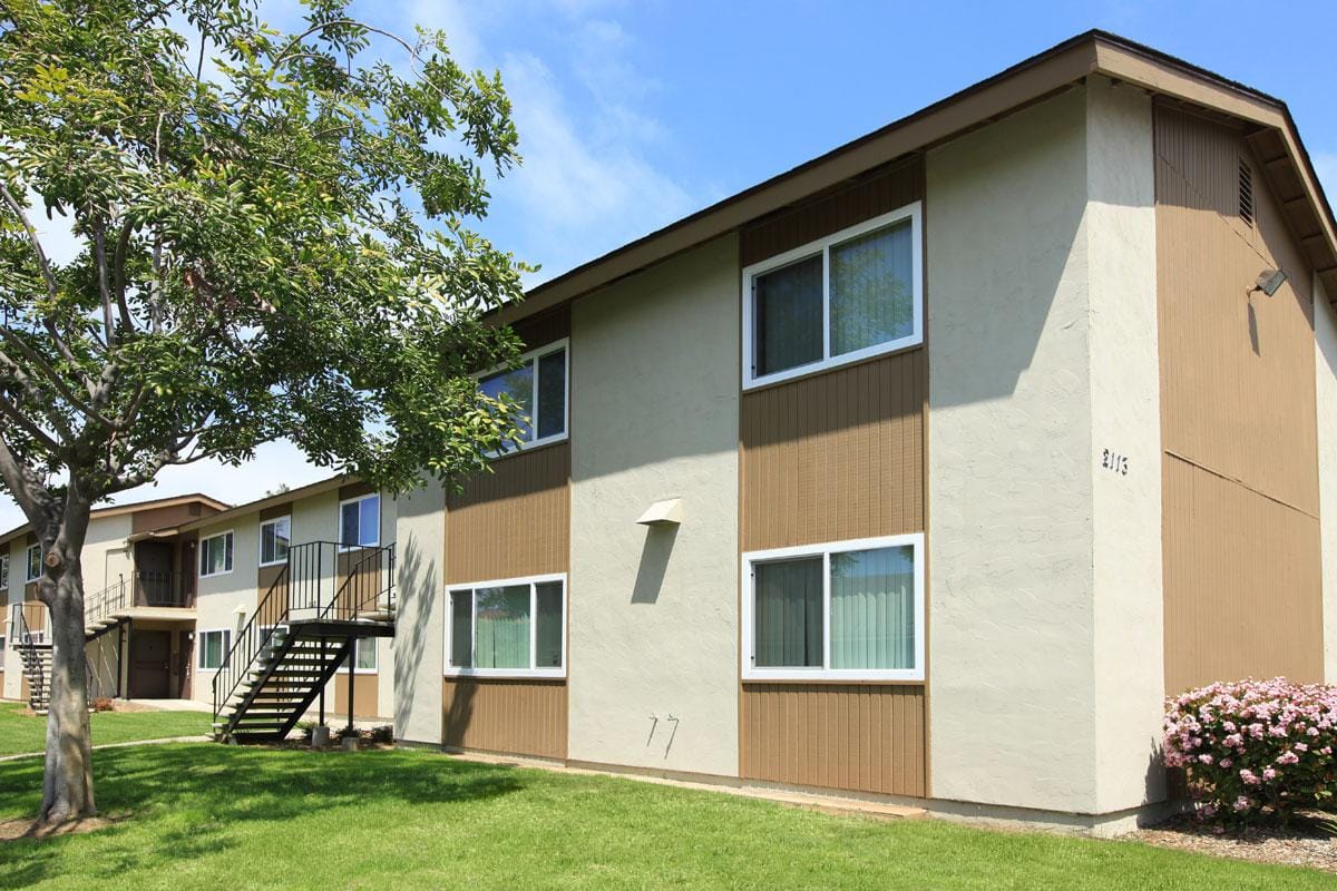 Exterior of a two story apartment building with green grass outside, a tree, and a sidewalk.