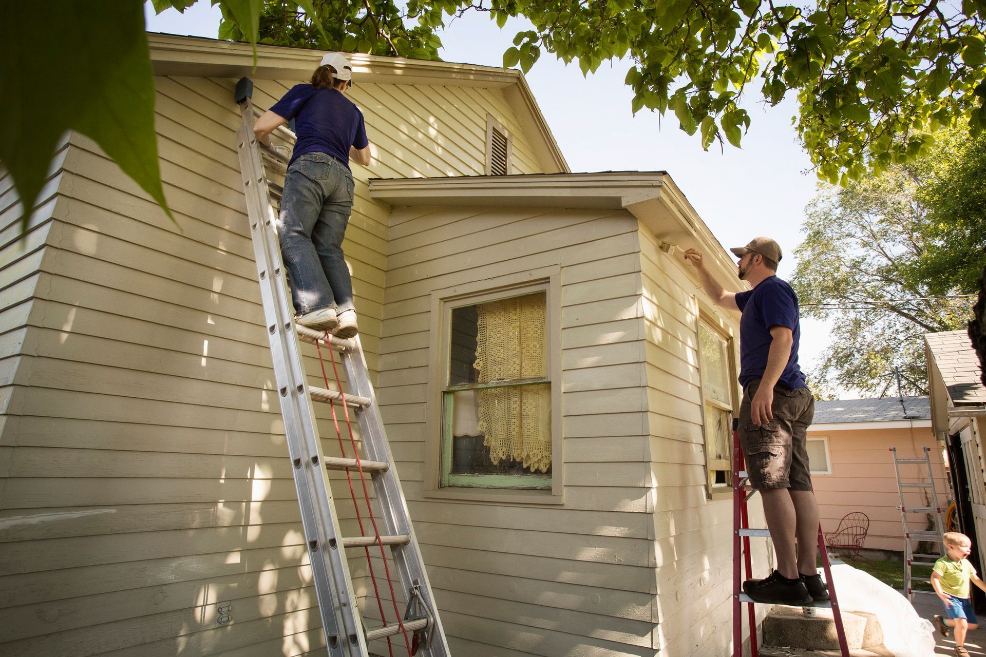 Two men on ladders repair siding on a light-colored house, sunny outdoor setting.