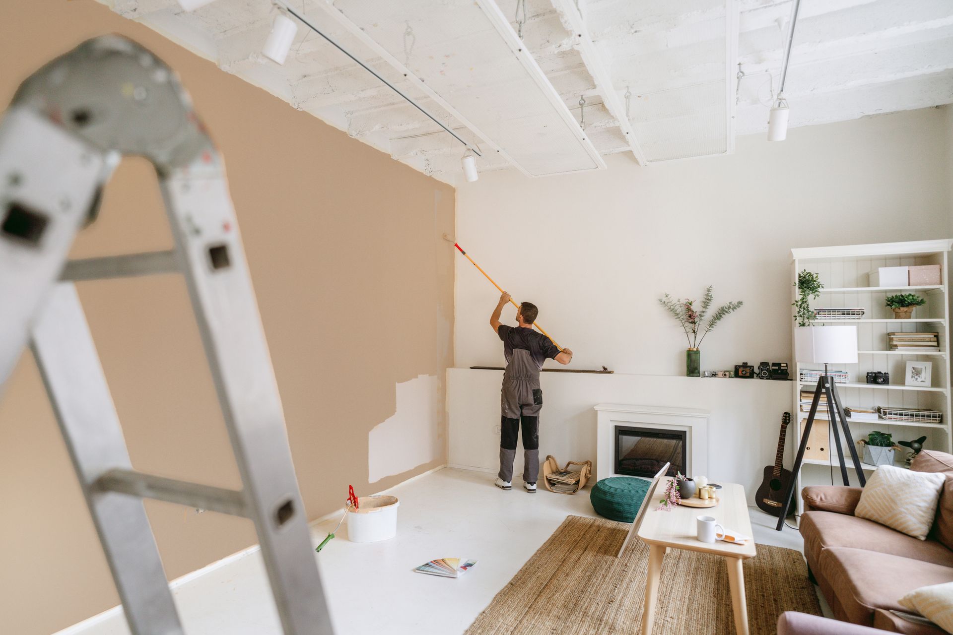Person painting a wall tan, using a roller. Ladder in view, living room setting.