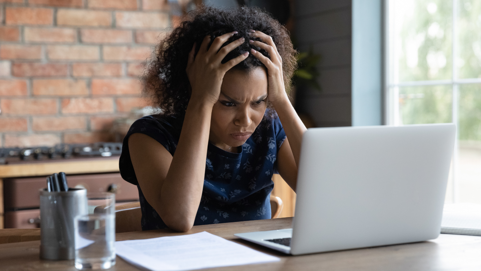 A woman is sitting at a desk confused with her hands on her head in front of a laptop computer.