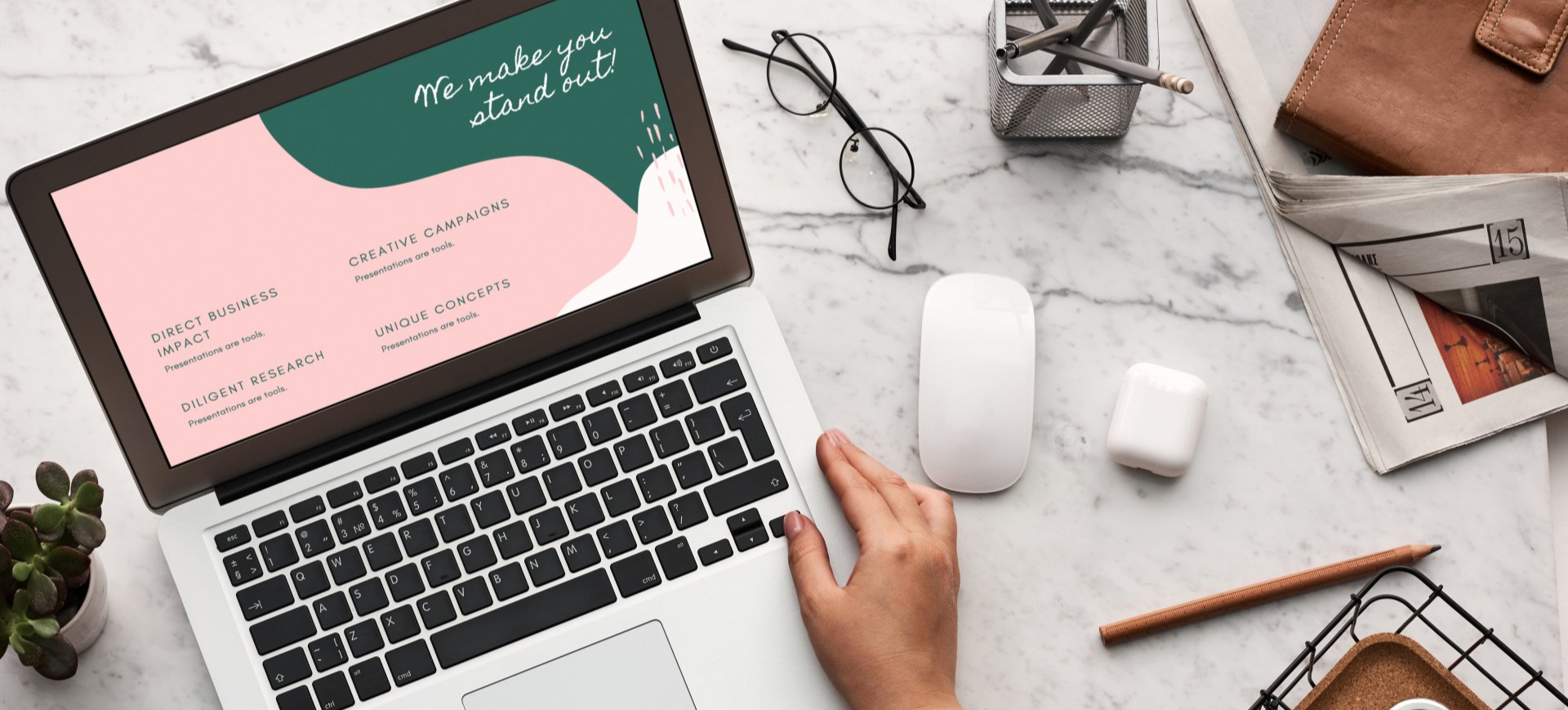Laptop on a marble surface with hands typing. Office supplies, glasses, and plants surround.