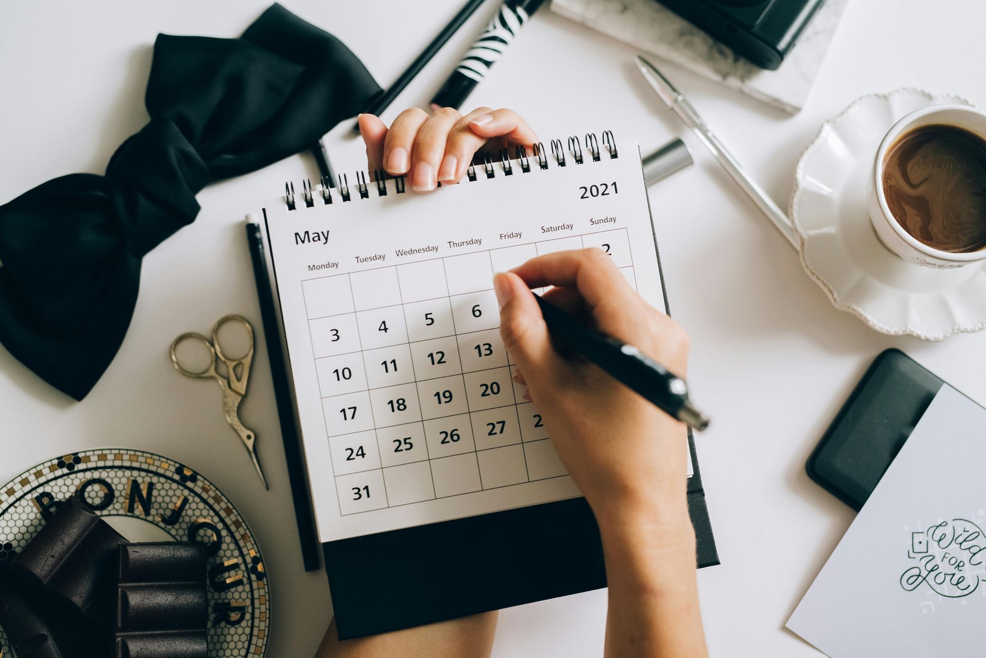 Person writing on a calendar, May 2021, on a white desk with coffee, bow tie, and other office supplies.