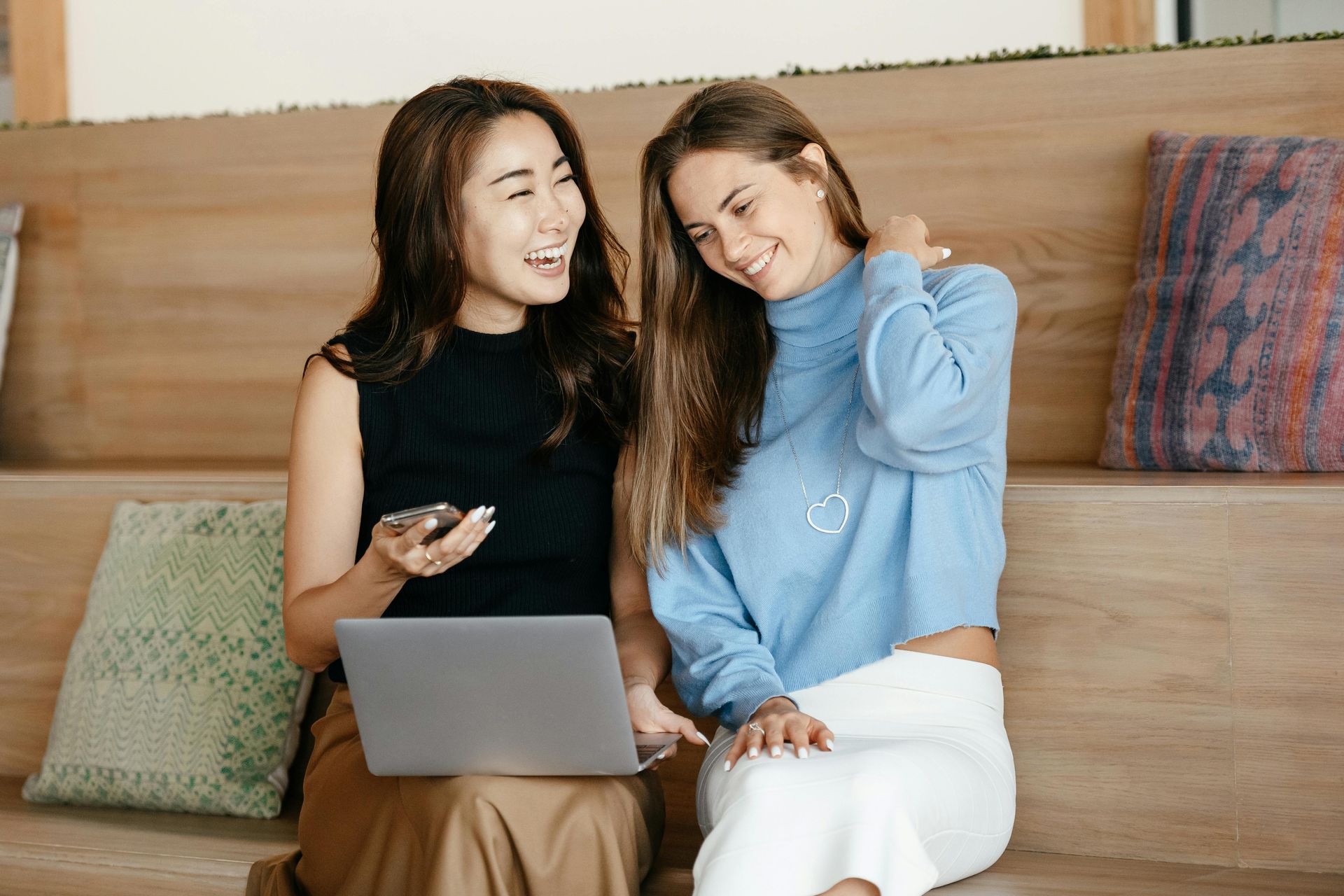 Two women sitting together on a couch, smiling about the content they are looking at on a laptop.
