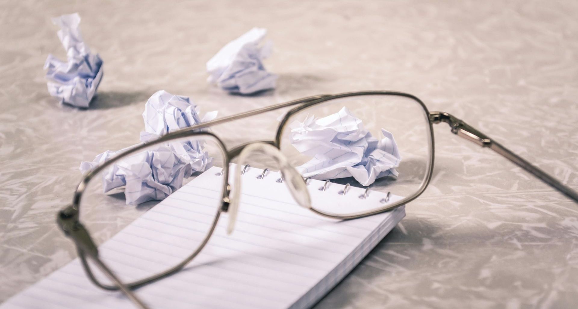 Eveglasses sitting on an empty notepad, with 5 crumpled-up balls of paper.