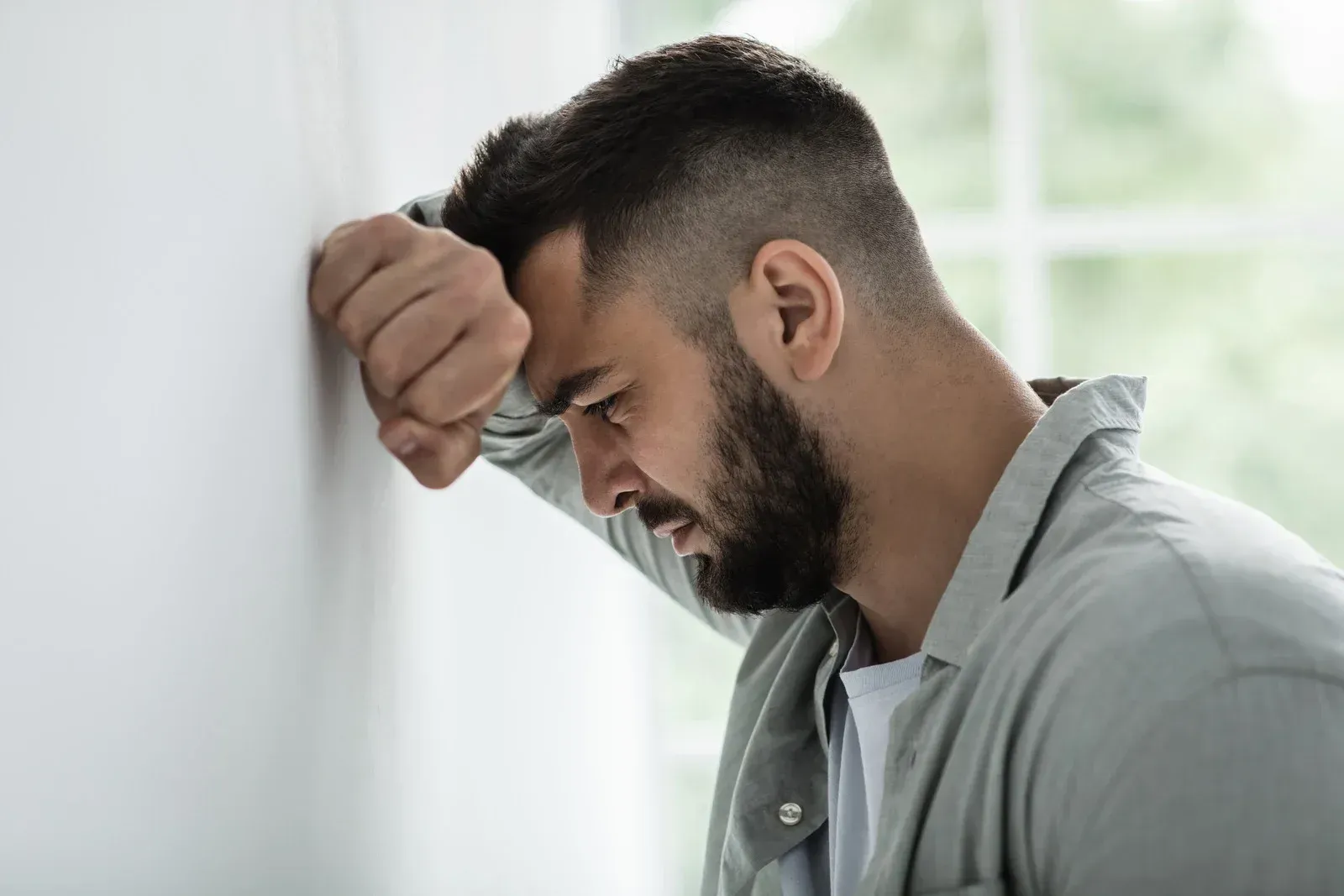 Man with beard presses head and fist against a white wall, appearing distressed.