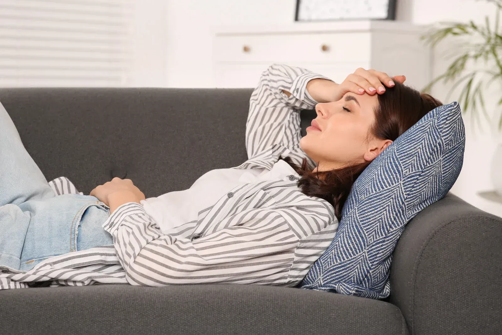 Woman with hand on forehead lies on gray couch, appearing unwell.