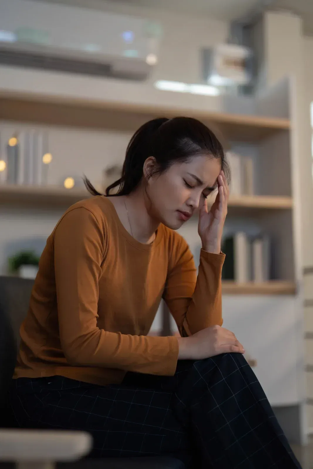 Woman with a headache, holding her head. She sits indoors, eyes closed, wearing orange shirt.