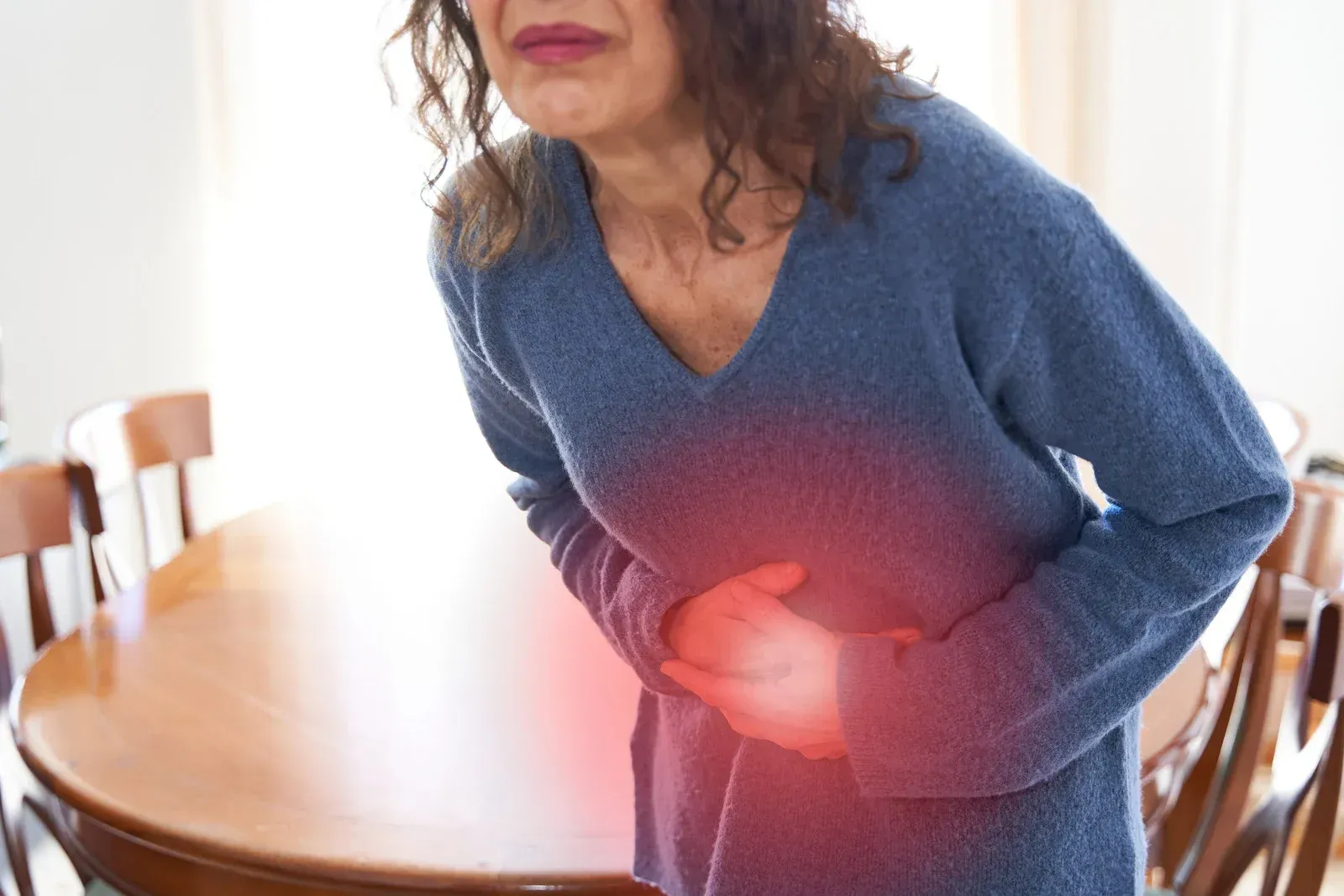 Woman holding stomach, appearing in pain, near a wooden table.