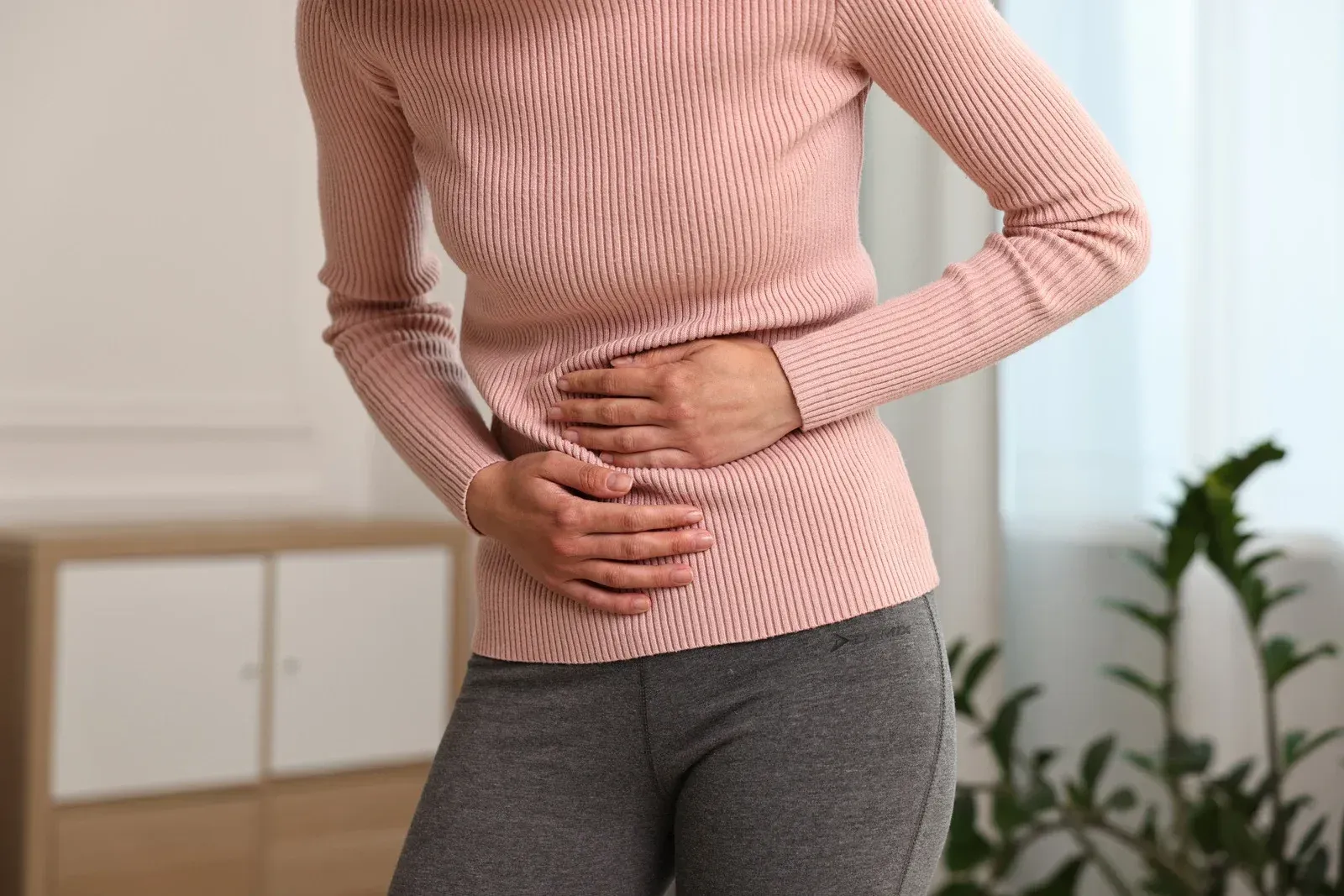 Woman in pink sweater and gray pants holding stomach, possibly in pain.