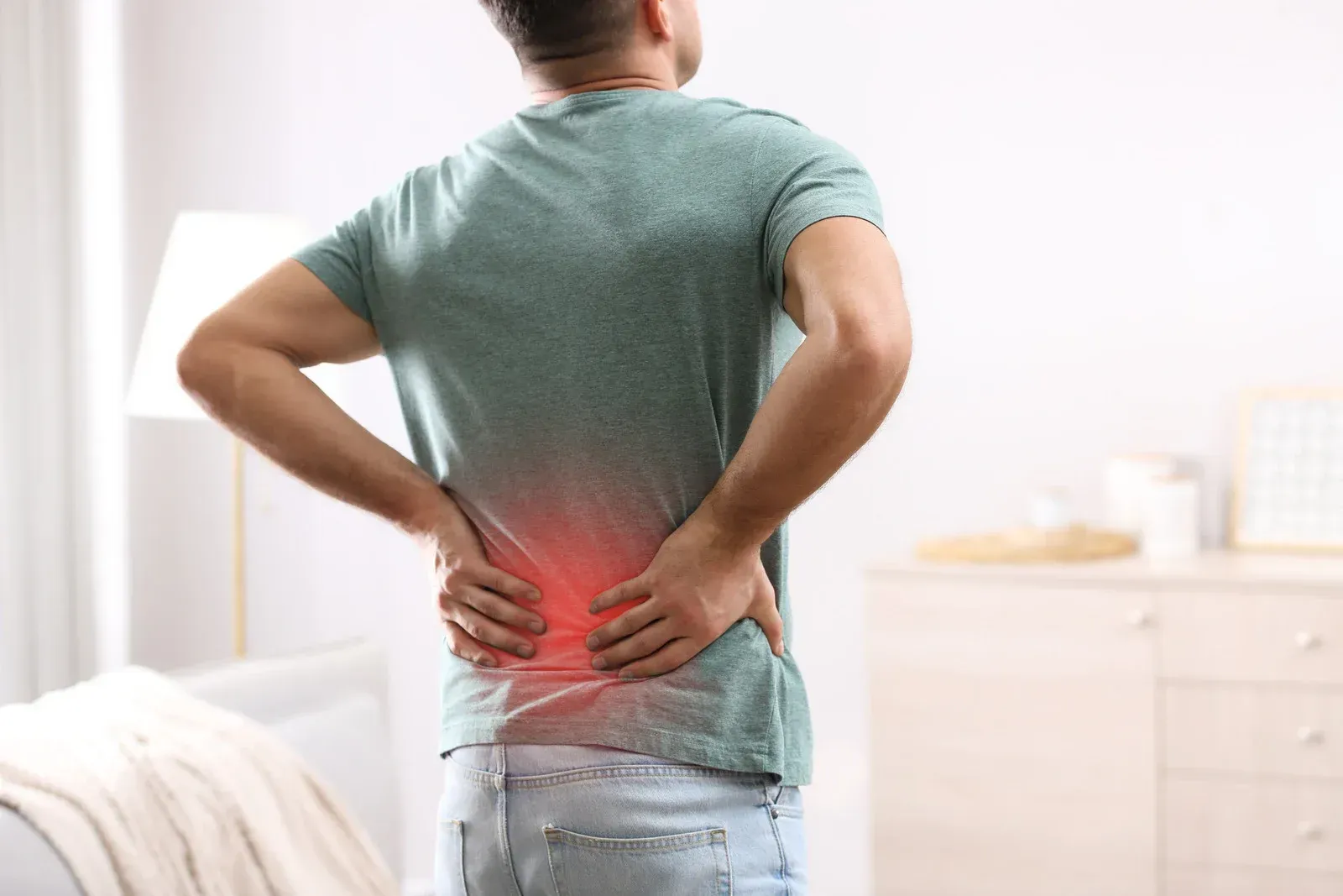 Man in a green shirt holding his back, indicating lower back pain, in a home setting.