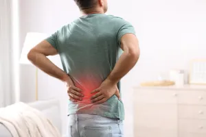 Man in a green shirt holding his back, indicating lower back pain, in a home setting.