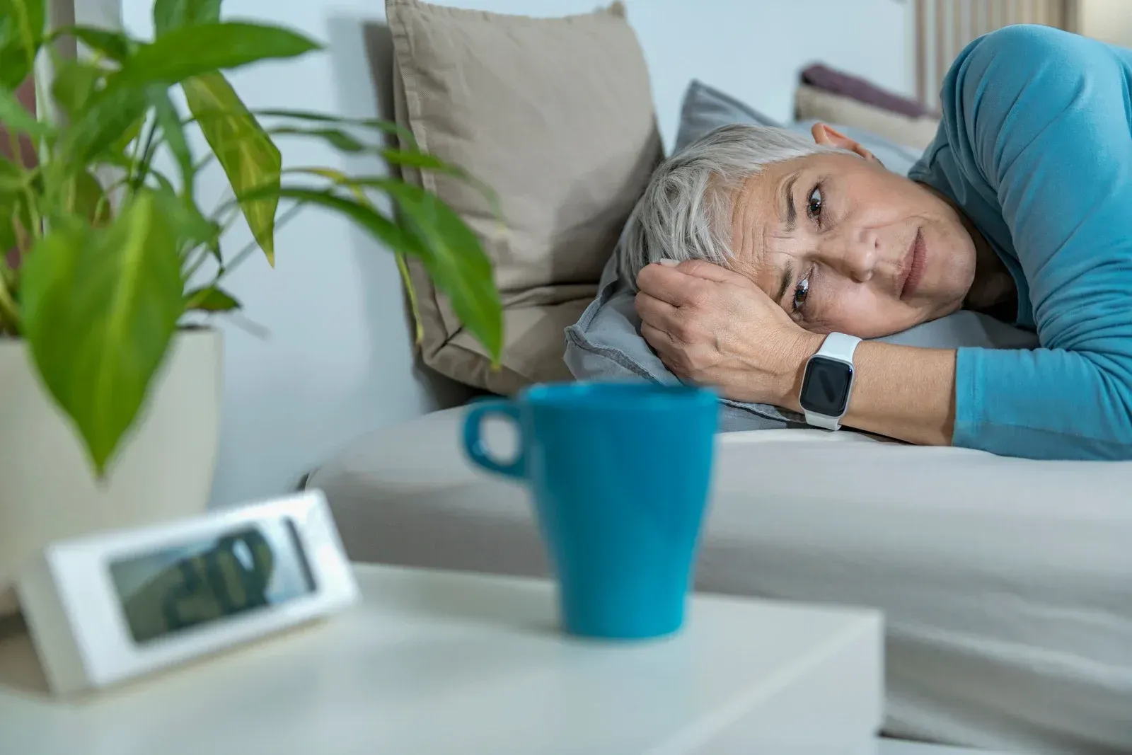 Woman in bed, wearing a watch, with a coffee cup and digital clock on a nightstand.