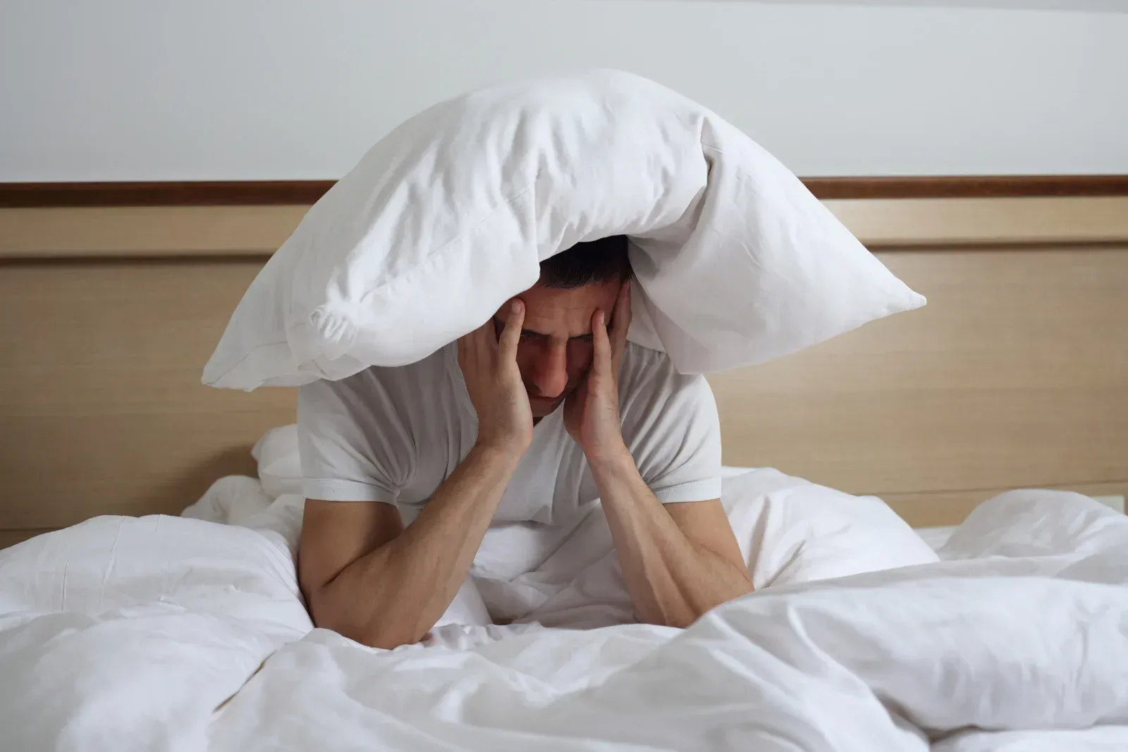 Man in bed with pillow over his head, hands on his temples, looking stressed.