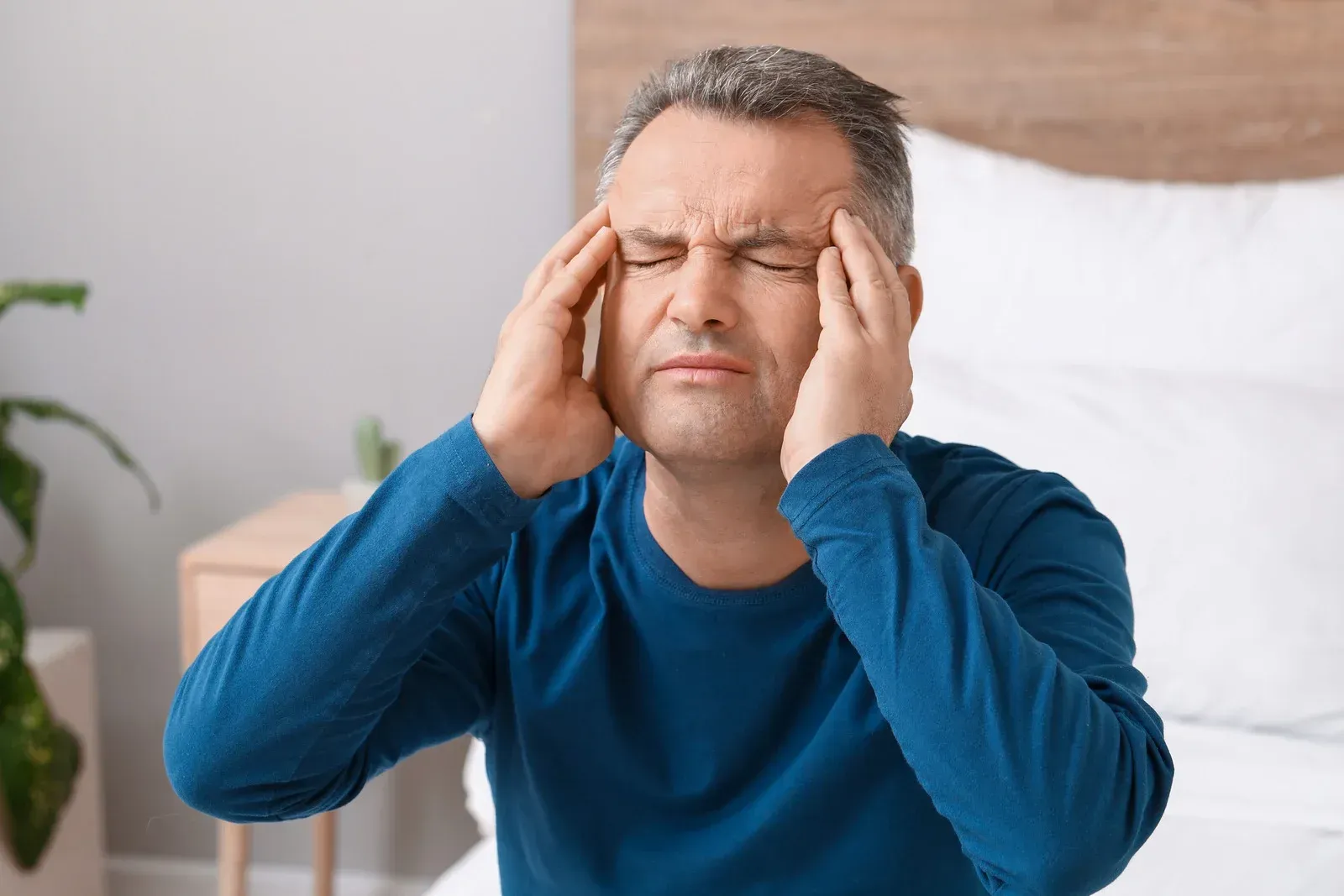 Man with gray hair in a blue shirt, holding his temples, eyes closed, appearing to have a headache, sitting on a bed.