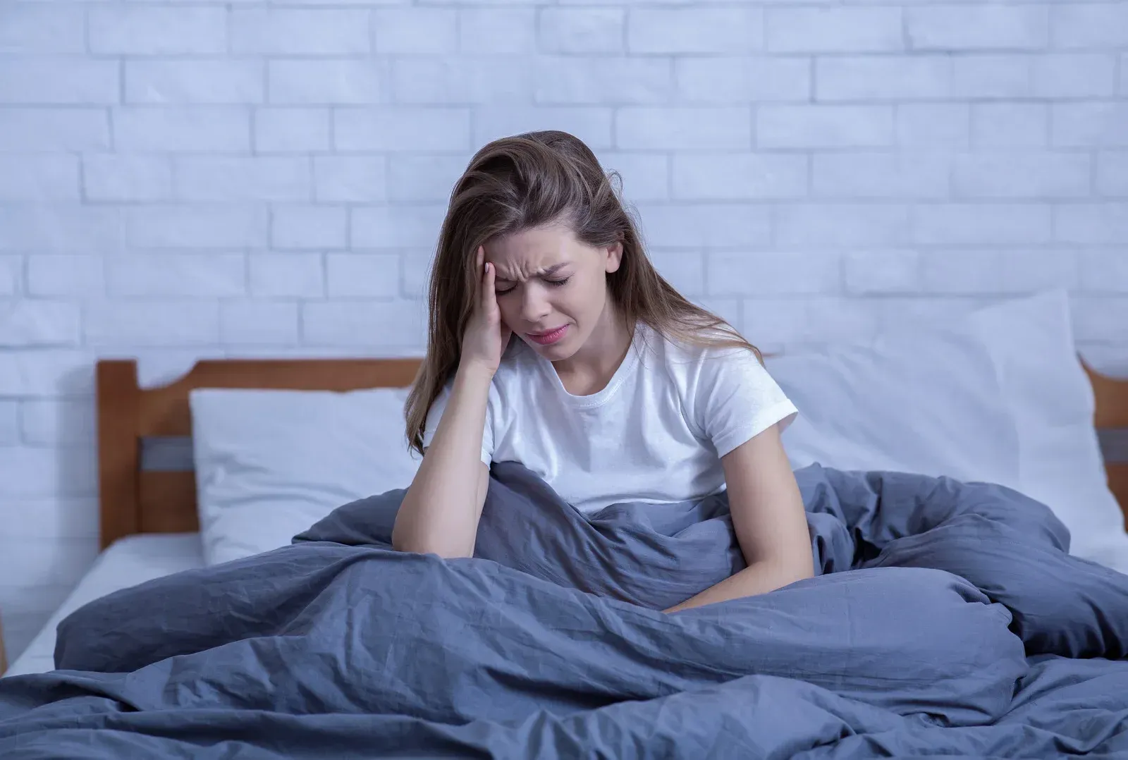 Woman with a headache sits in bed, holding her head. Dark gray comforter, white wall.