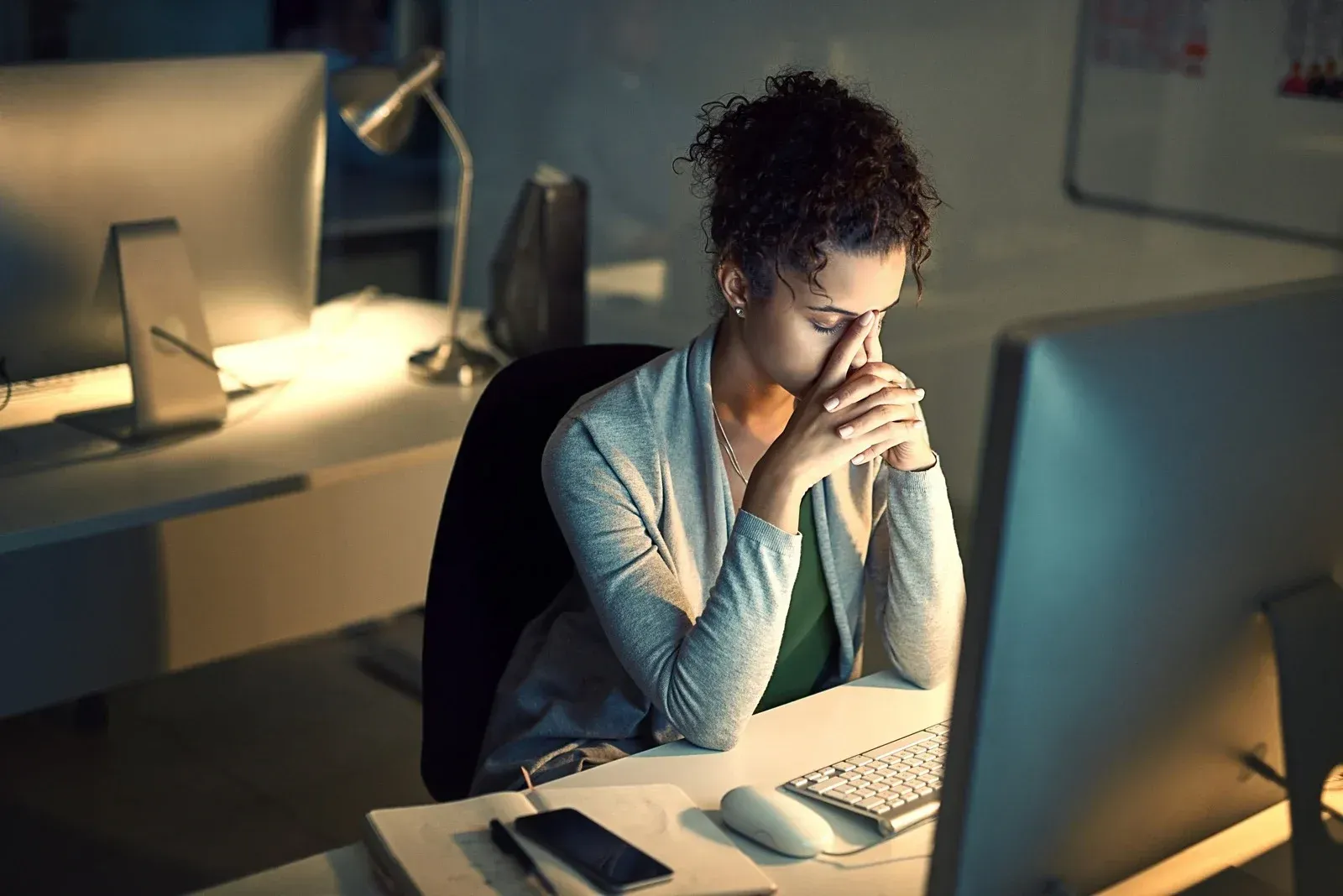 Woman stressed at a computer, hands on face in a dimly lit office.