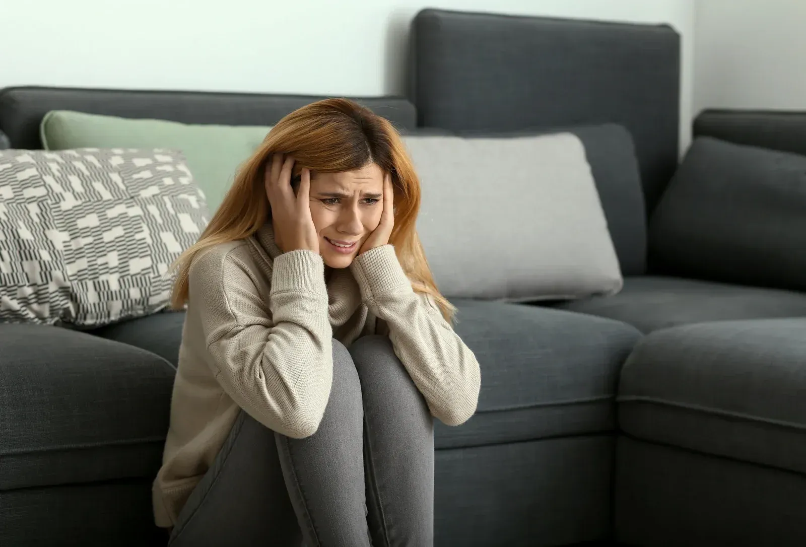Woman with hands covering ears, curled up on floor near sofa, looking distressed.
