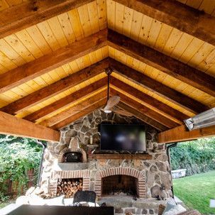 A wooden gazebo with a stone fireplace and a flat screen tv.