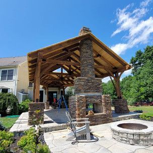 A wooden gazebo with a stone fireplace and a fire pit in front of a house.
