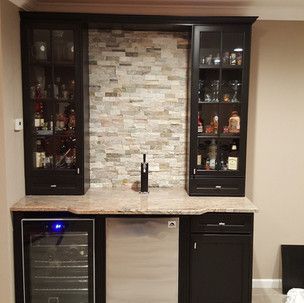 A kitchen with black cabinets , a stainless steel refrigerator , a sink , and a stone wall.