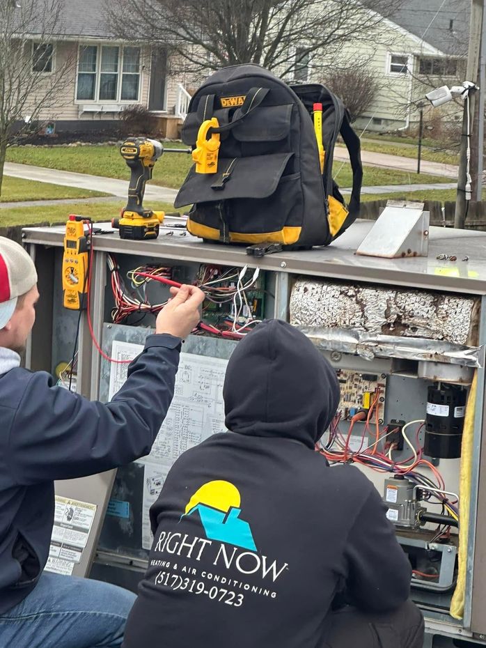 Two technicians in branded hoodies work on an opened HVAC unit outdoors, using tools and a multimeter.