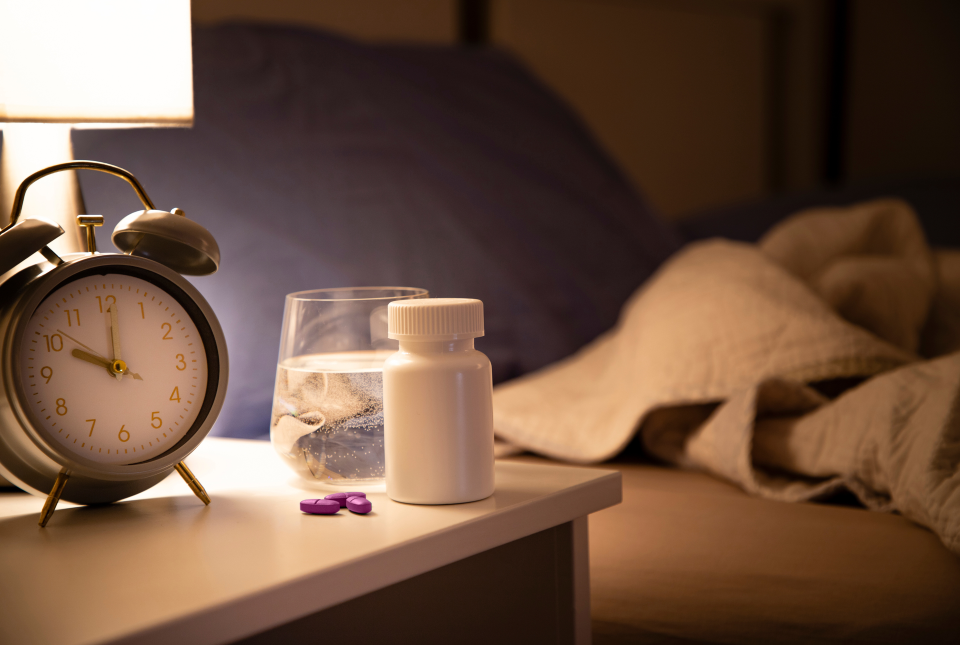 Alarm clock, pills, water glass, and medicine bottle on a nightstand next to a bed.