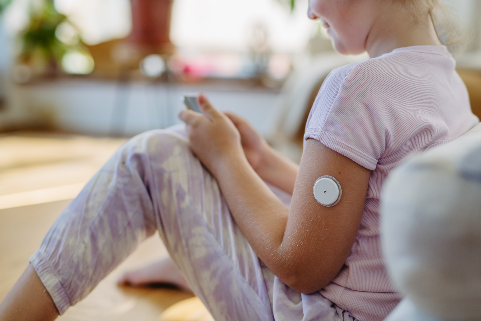 A little girl with a patch on her arm is sitting on the floor using a cell phone.