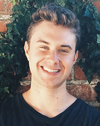 A young man in a black shirt is smiling in front of a brick wall.