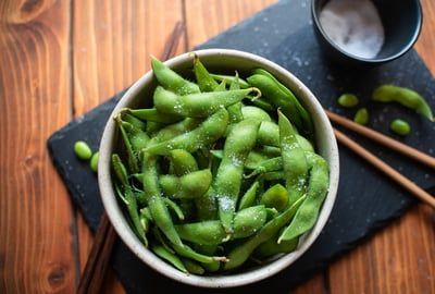 A bowl of edamame beans with chopsticks on a wooden table.