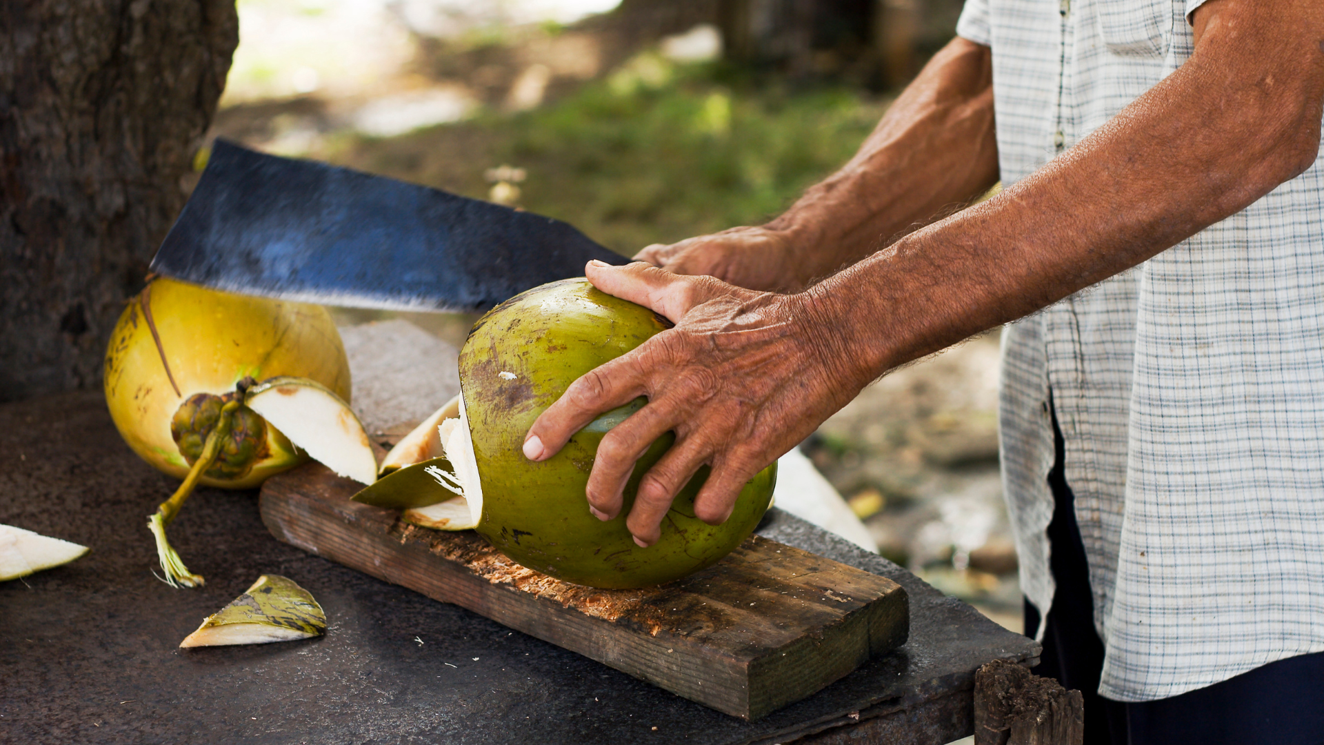 A man is cutting a coconut with a machete on a cutting board.