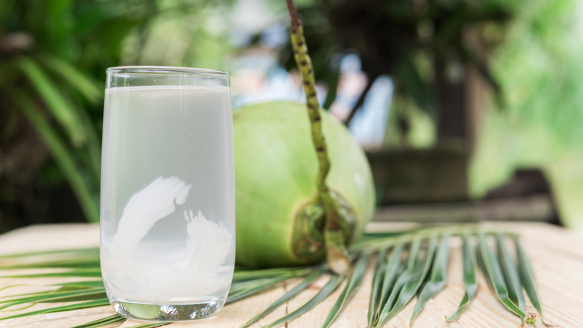 A glass of coconut water is sitting on a wooden table next to a coconut.