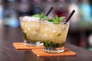 Two mojito cocktails with mint leaves and straws on a wooden coaster on a bar.