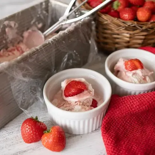 Two bowls of ice cream with strawberries on top are on a table next to a basket of strawberries.