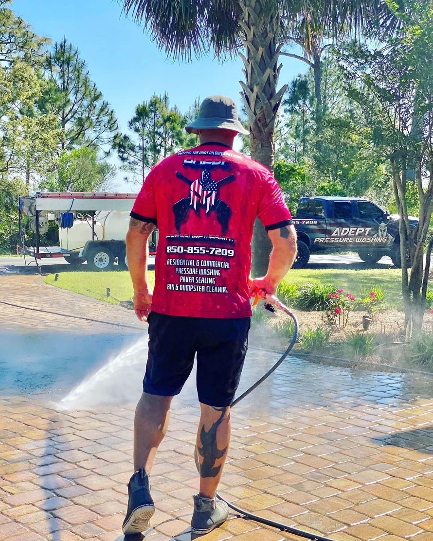 A person in a red uniform power washes a brick driveway next to a service vehicle on a sunny day.