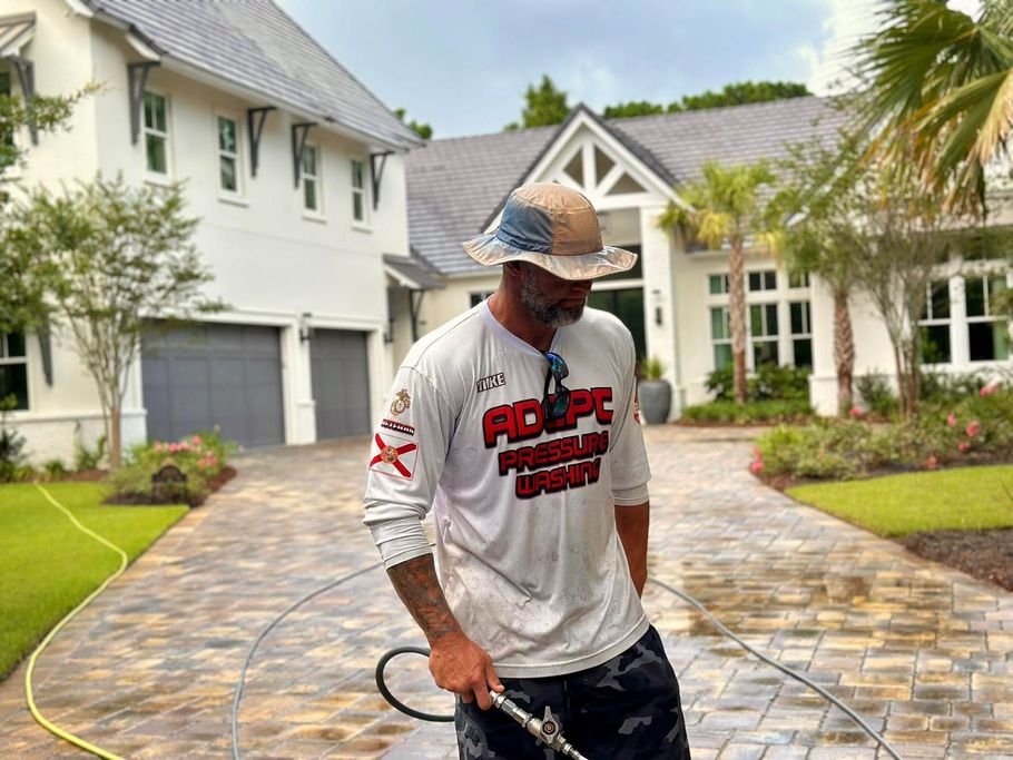 A person in a sun hat and logo shirt uses a pressure washer to clean a paved driveway in front of a white house.