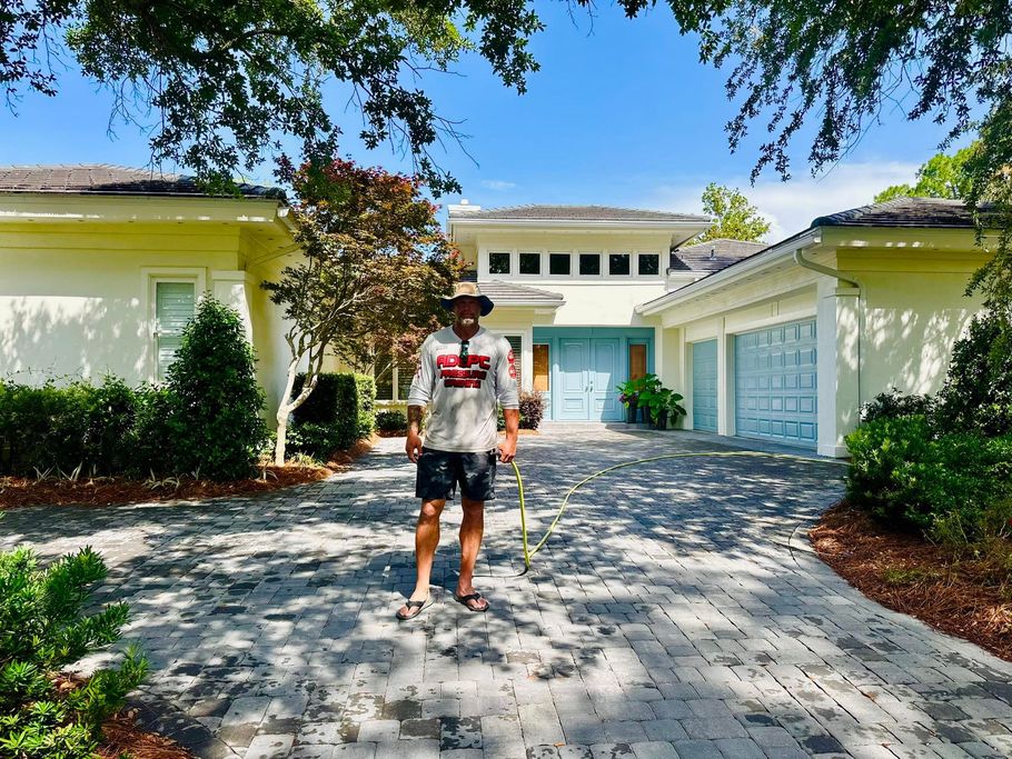 A person standing on a patterned paver driveway in front of a light-colored house with a blue door and garage doors.