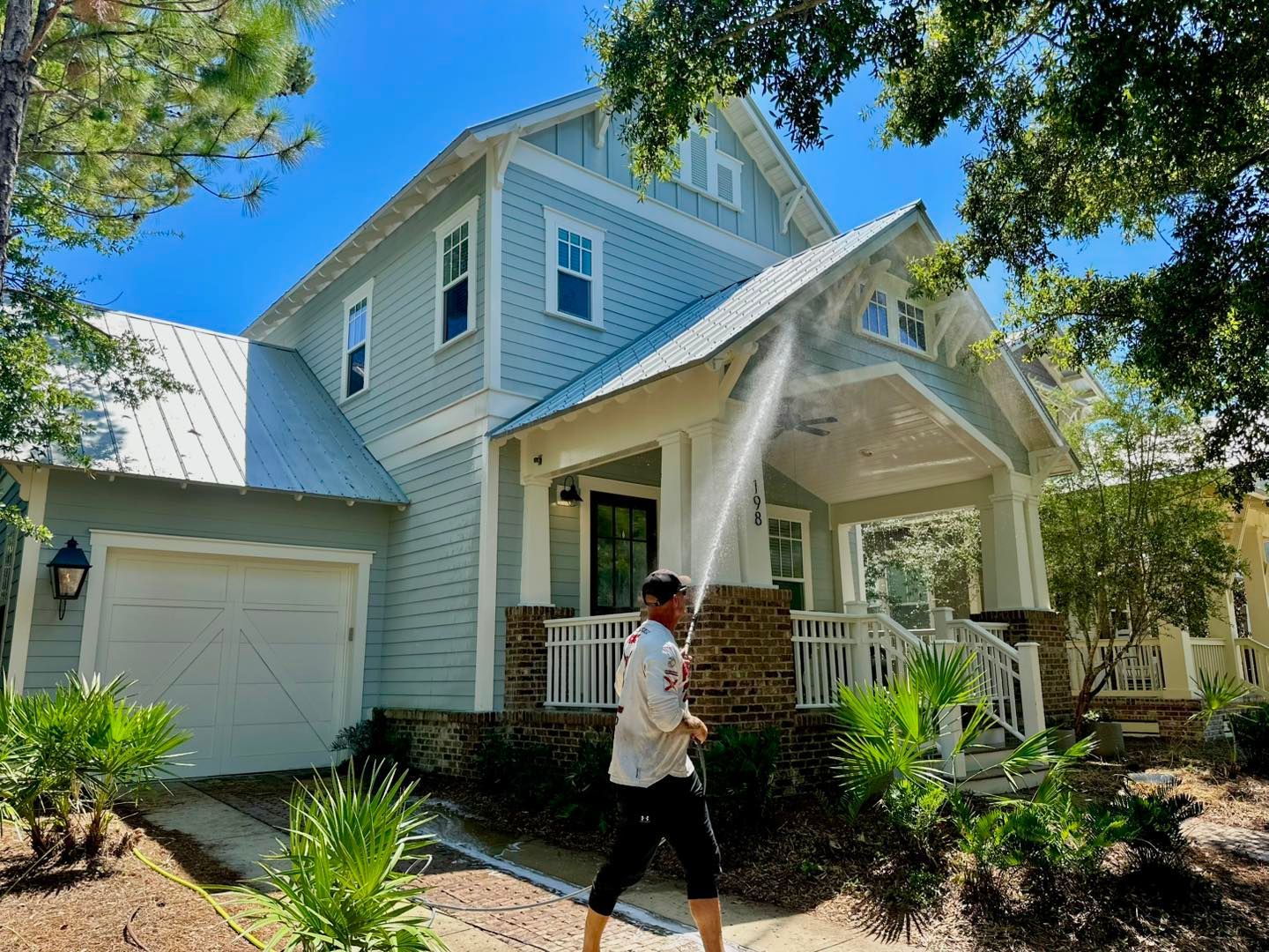 A person uses a pressure washer to clean the exterior of a light blue two-story house on a sunny day.