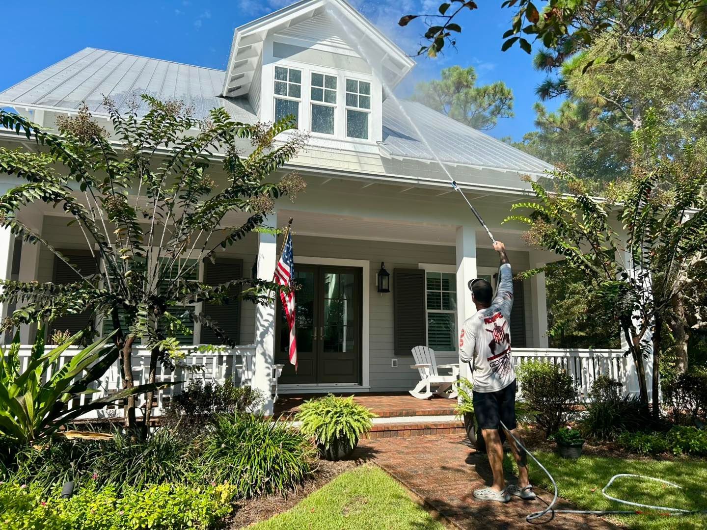 A person uses a pressure washer to clean the metal roof of a light-colored house with a front porch and American flag.