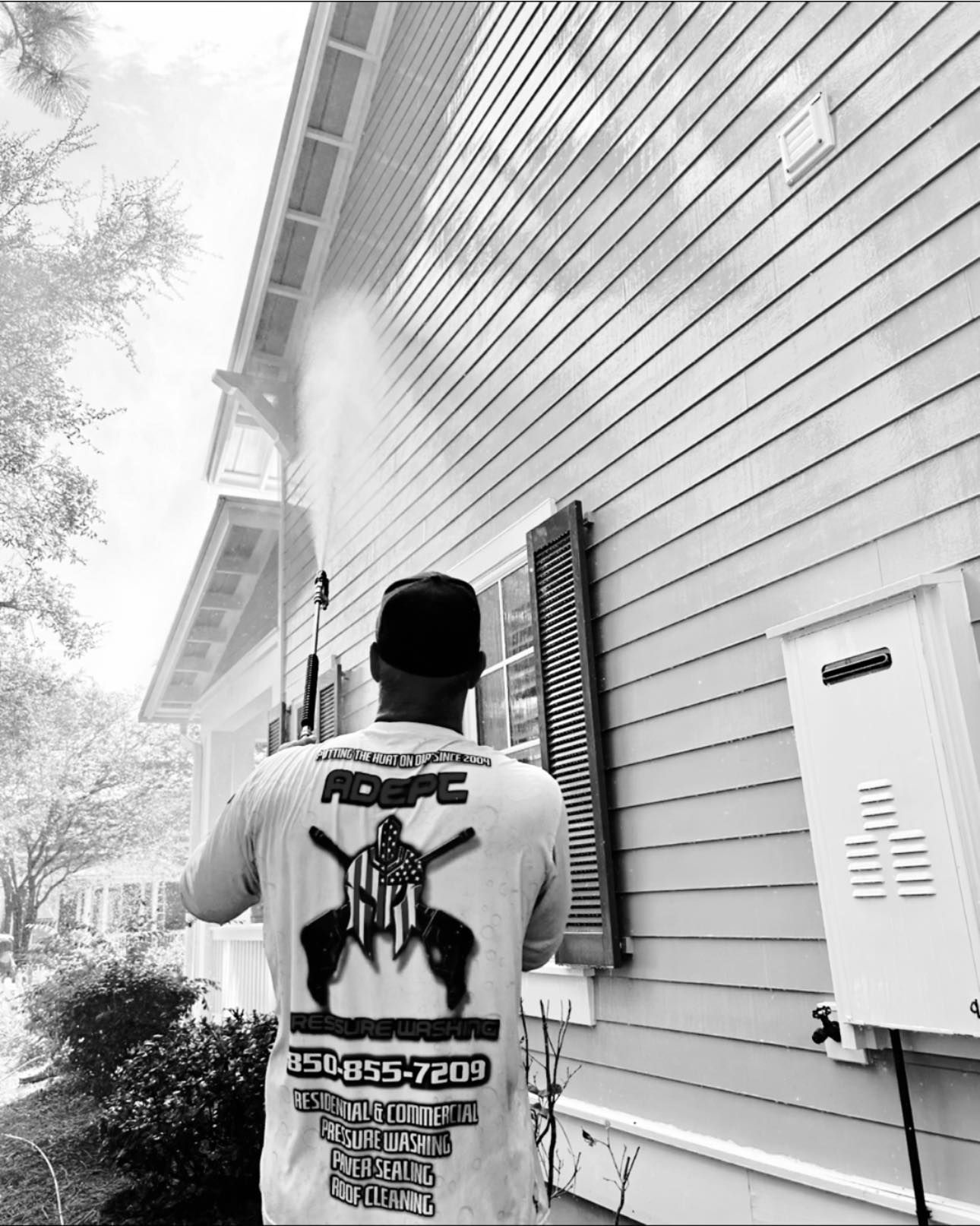 A person in a logo t-shirt uses a high-pressure spray wand to clean the exterior siding of a residential house.