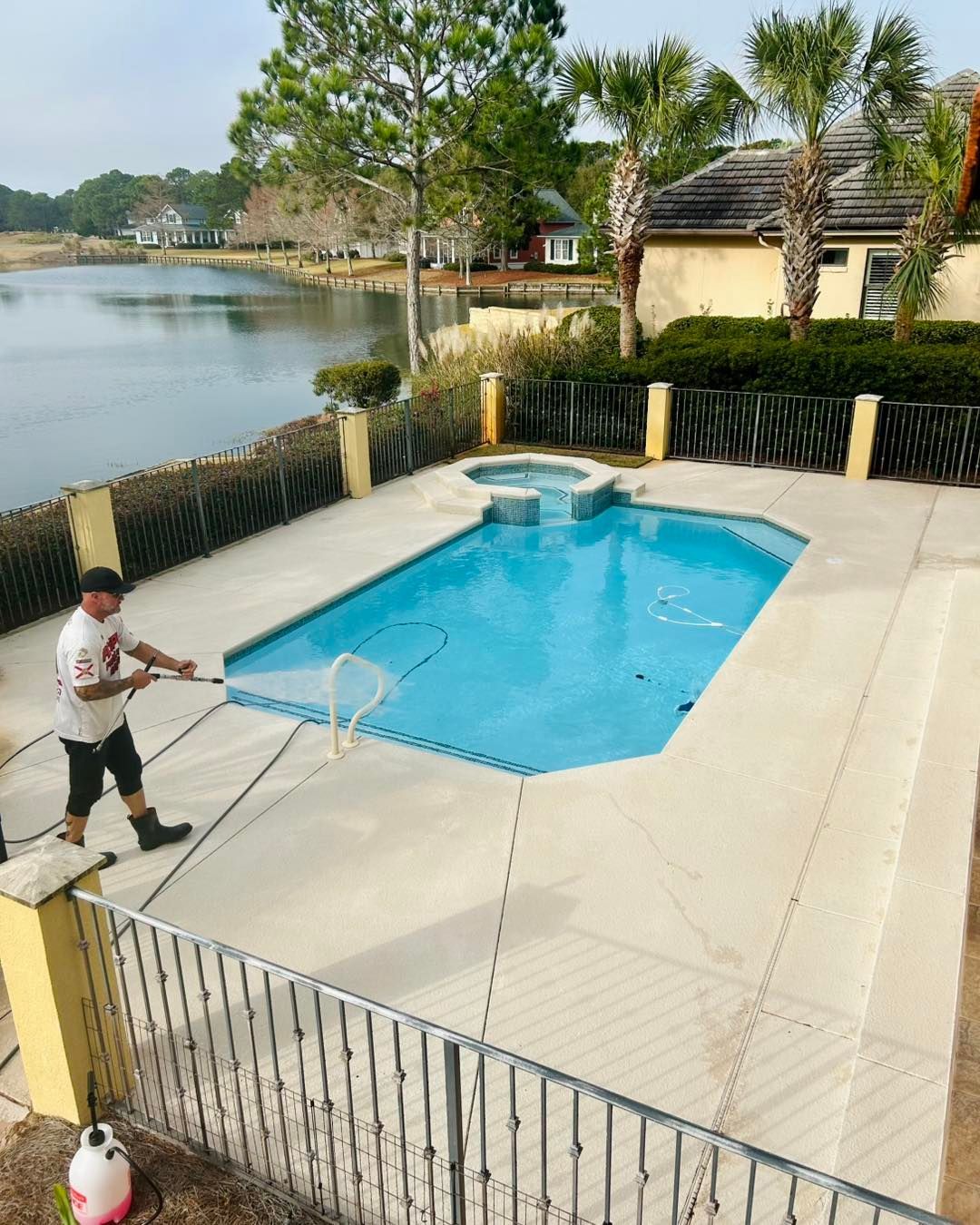 A person uses a net to clean a rectangular swimming pool with an attached spa, located by a lake on a sunny day.