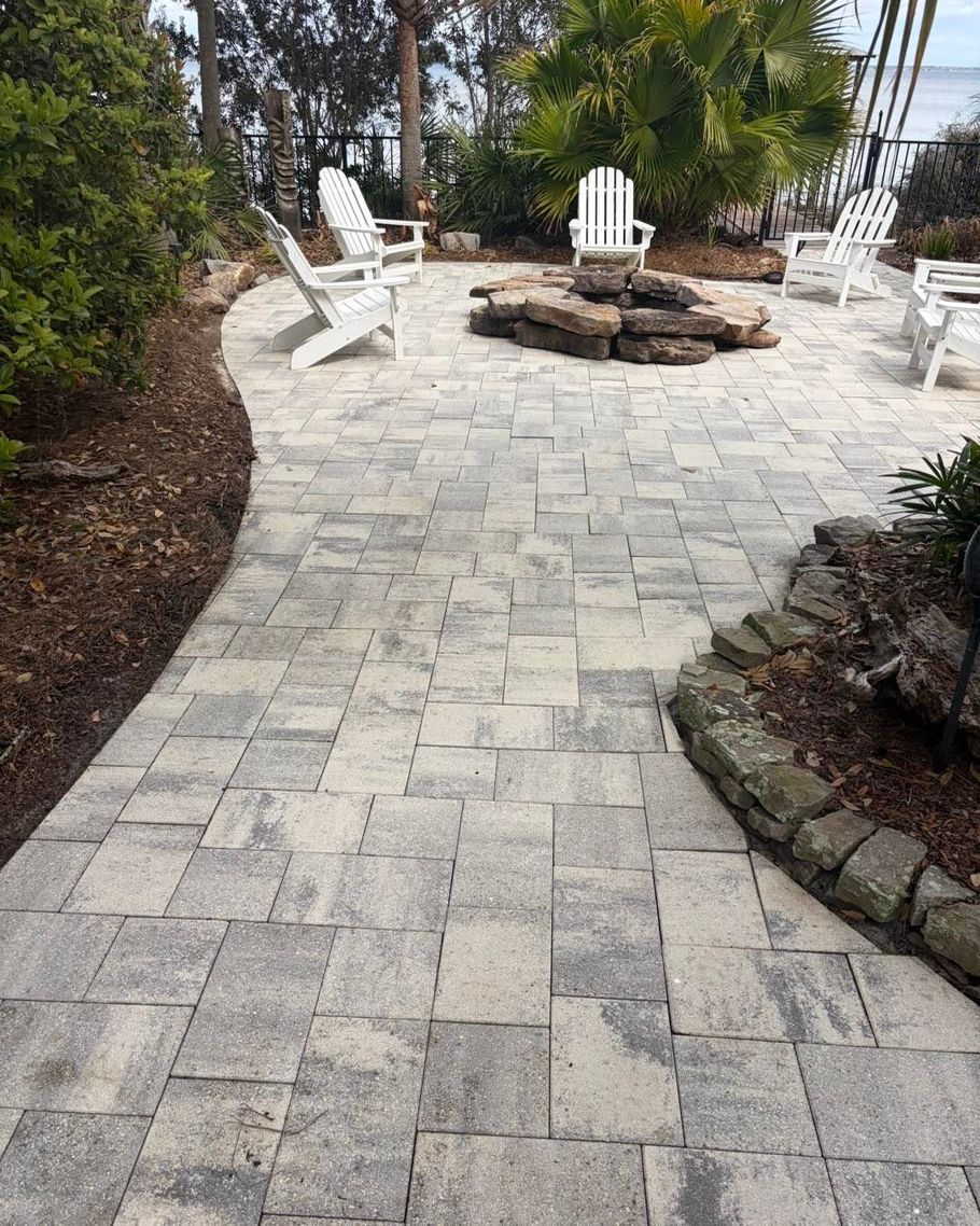 A patio with light-colored pavers, a stone fire pit, and several white Adirondack chairs in a landscaped backyard.