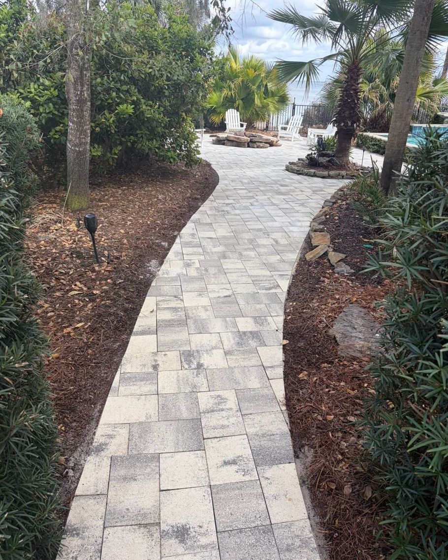 A light-colored stone paver path winds through a landscaped garden toward a distant patio and palm trees.