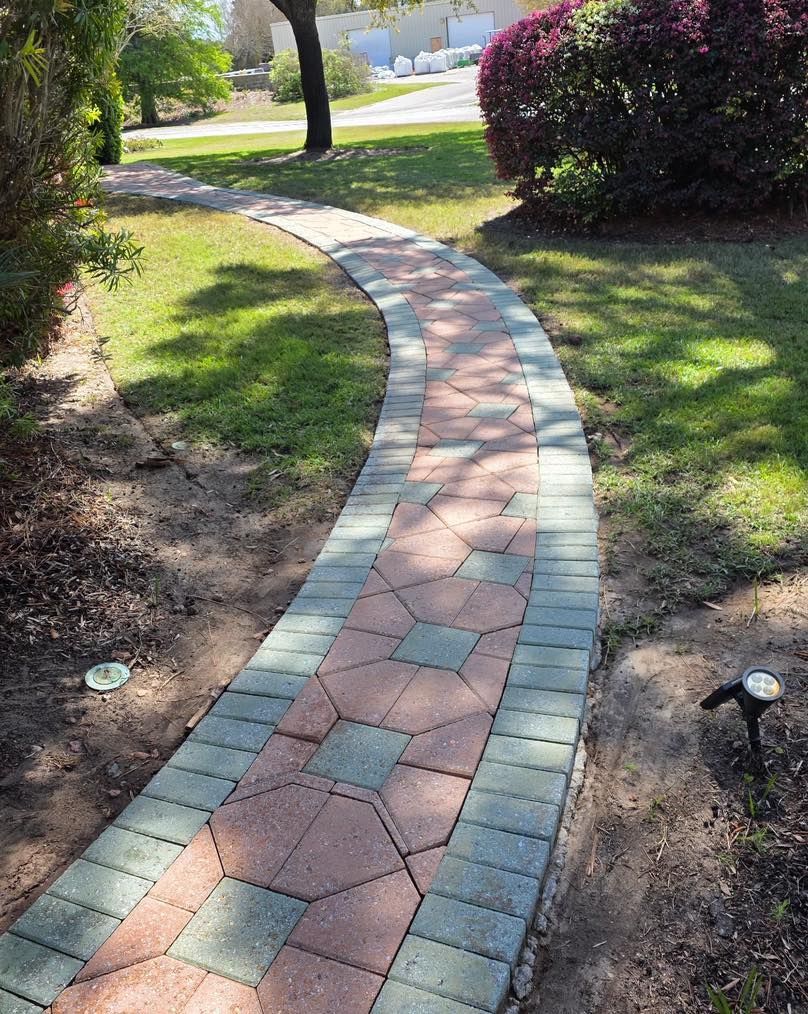A curved stone paver walkway with a geometric red and grey tile pattern, flanked by grass and landscape lighting.