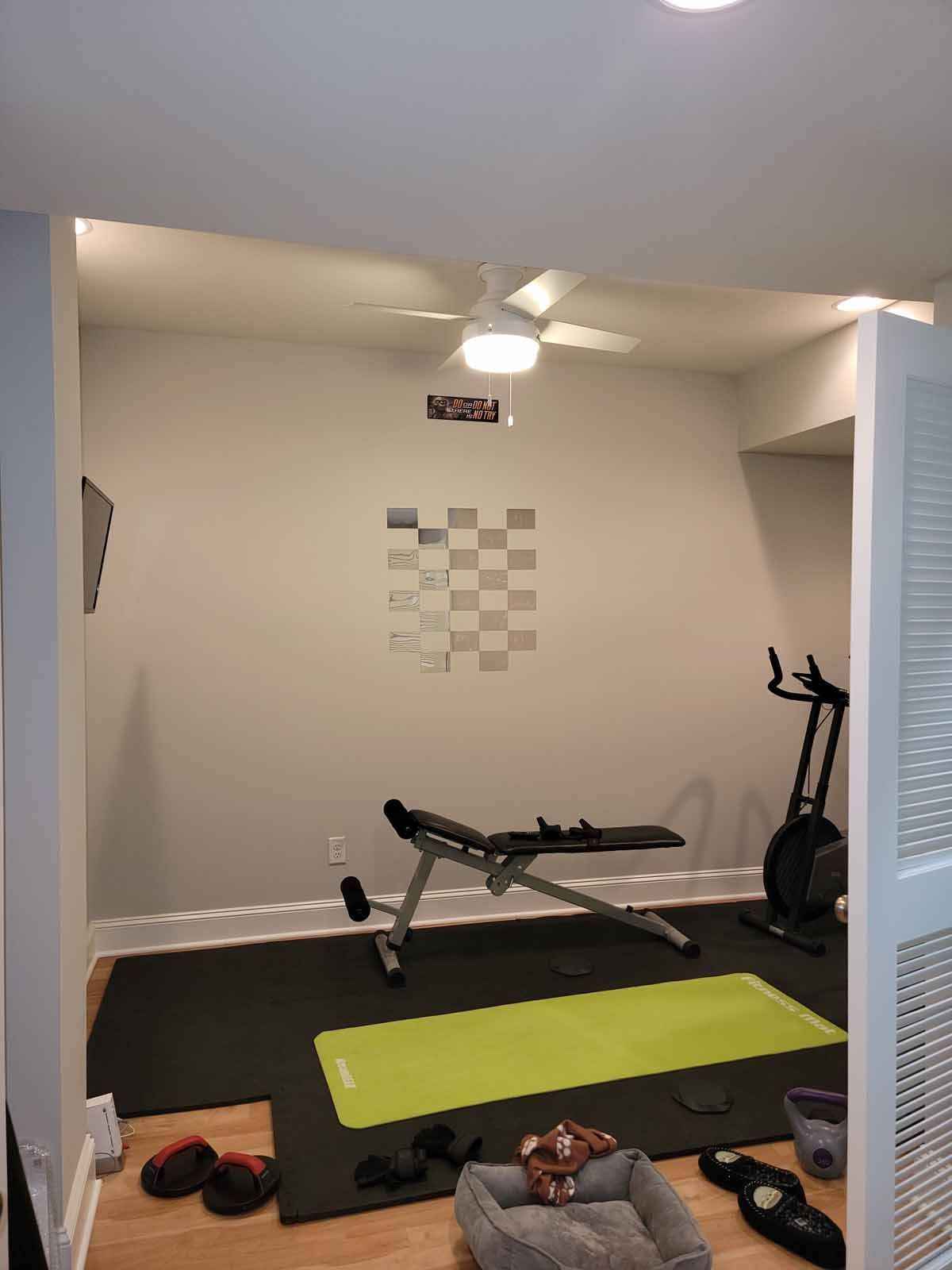 A gym with a ceiling fan , a yoga mat , a bench , and a dog bed.
