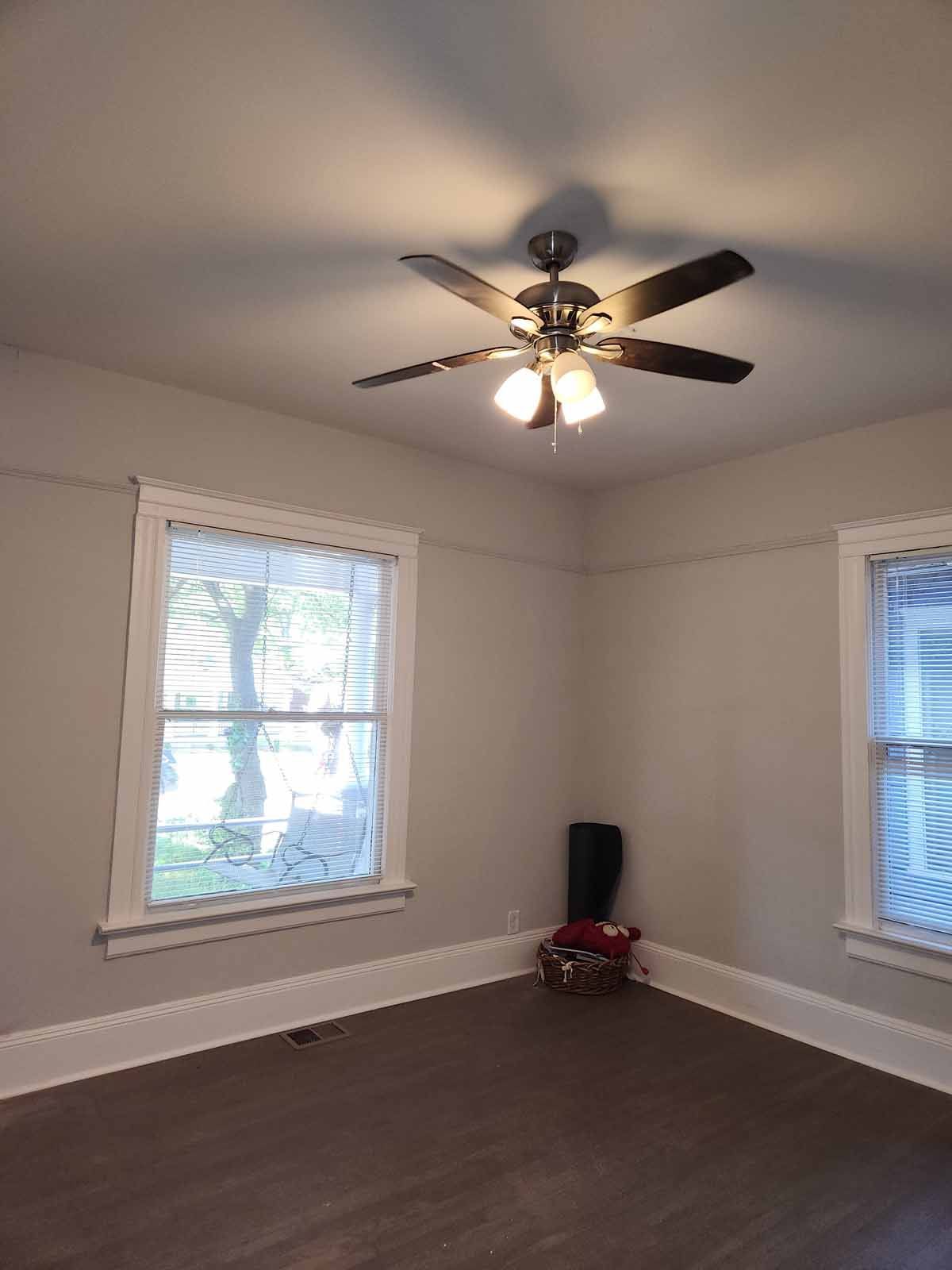 An empty room with a ceiling fan and two windows.