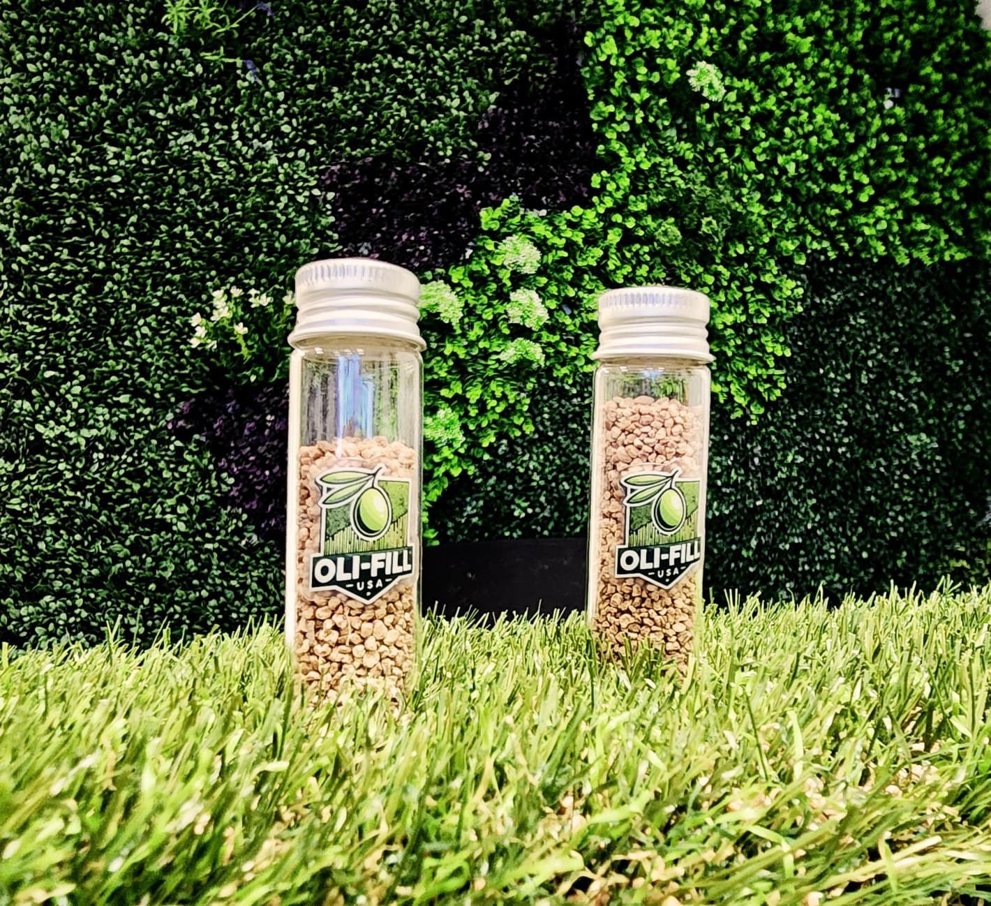 Two clear glass vials with seed-like granules stand on artificial grass against a green wall; the label reads 