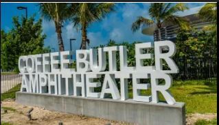 White sign for Coffee Butler Amphitheater. Green grass, palm trees, and blue sky in background.