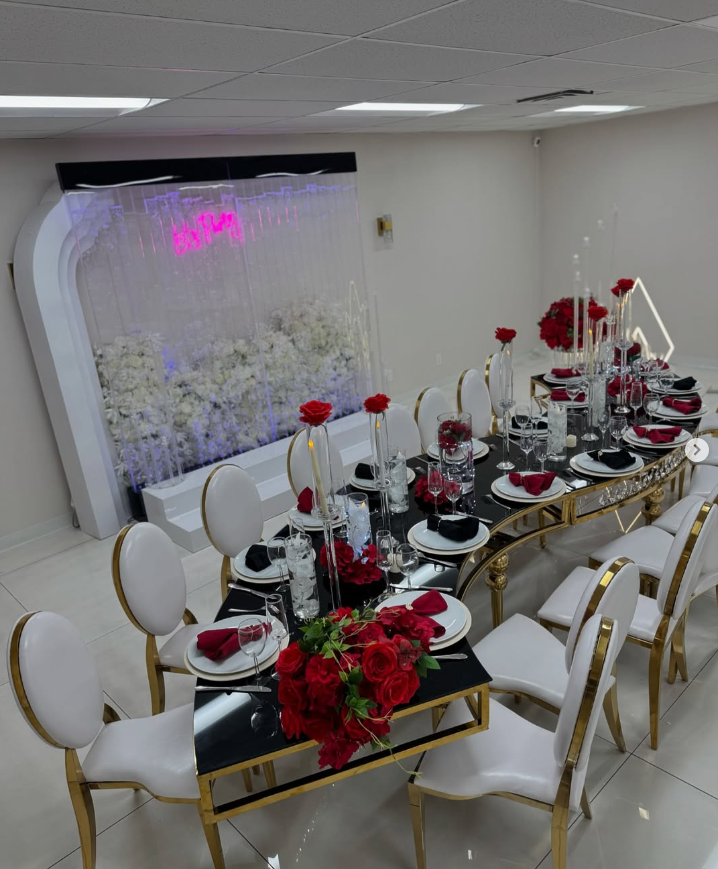 A long dining table with white chairs and red flowers on it.